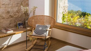 Una silla mecedora de mimbre con un cojín geométrico, un florero con flores blancas y vista al mar a través de una ventana.