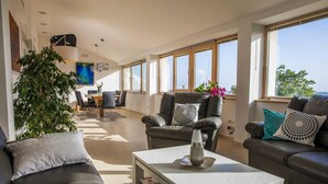 Una sala de estar moderna con un sofá, un sillón y una mesa de centro. Hay un comedor con una mesa y sillas. La habitación tiene ventanas grandes y una planta.