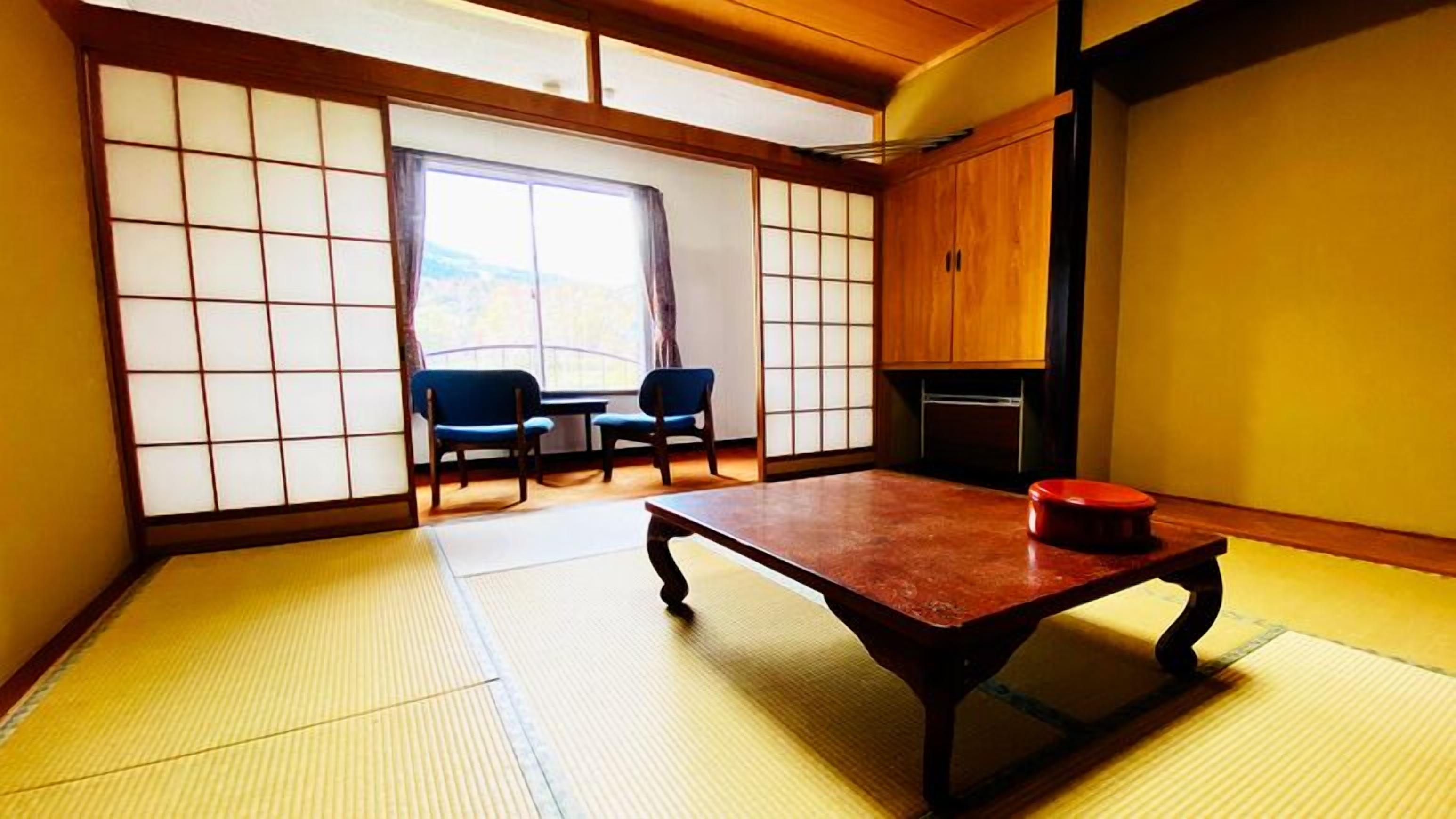 Una habitación tradicional japonesa con suelo de tatami, una mesa baja, dos sillas y puertas correderas shoji.