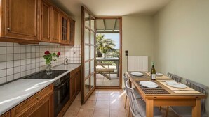 A kitchen with wooden cabinets, a tiled backsplash, a dining table set for two, and a view of a patio with a palm tree. - Villa 15  Elegant Private Pool Family (Rethymno)