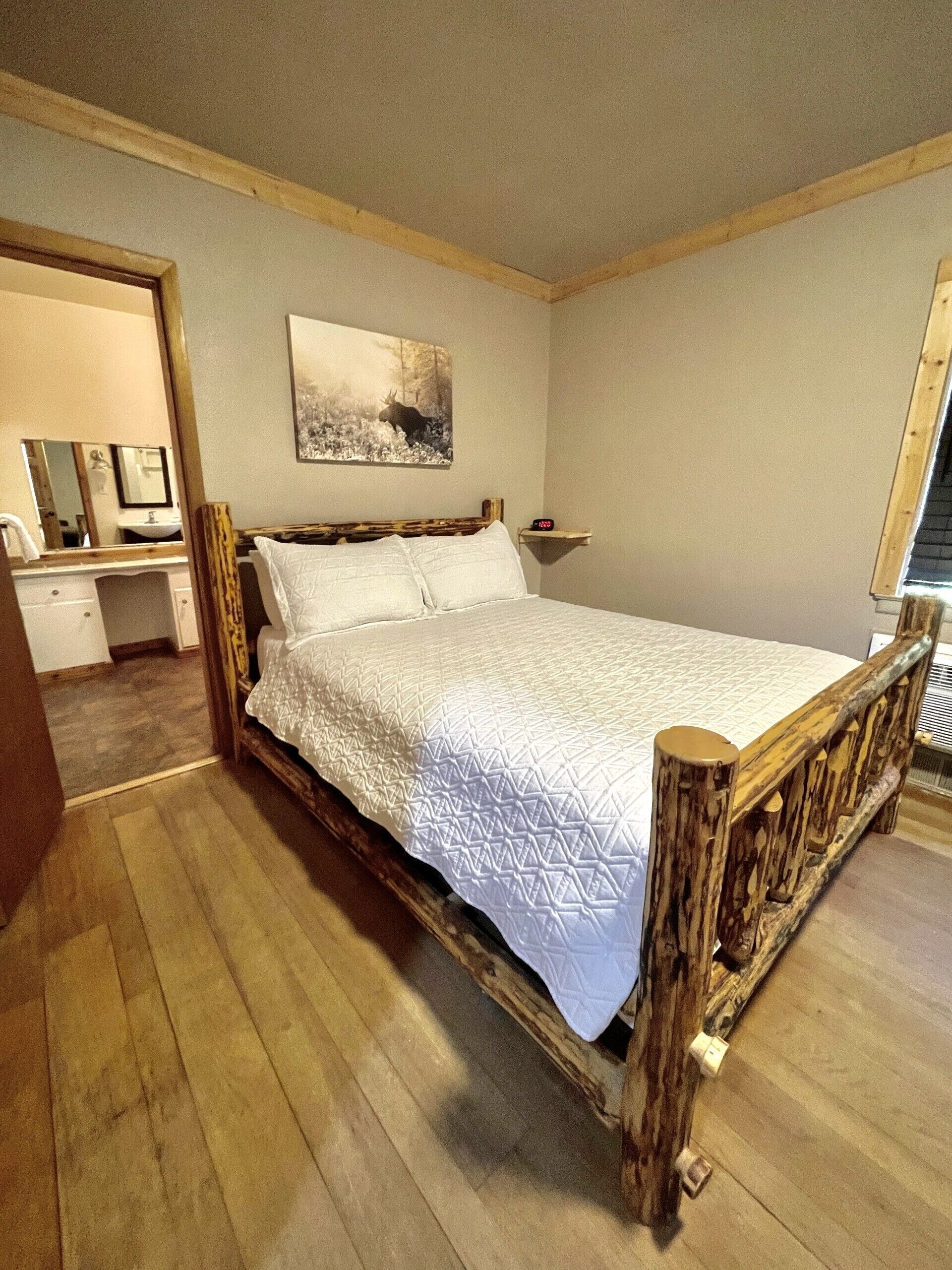 A bedroom with a wooden bed frame, a white quilted bedspread, a mounted picture of a moose, and a wooden headboard.