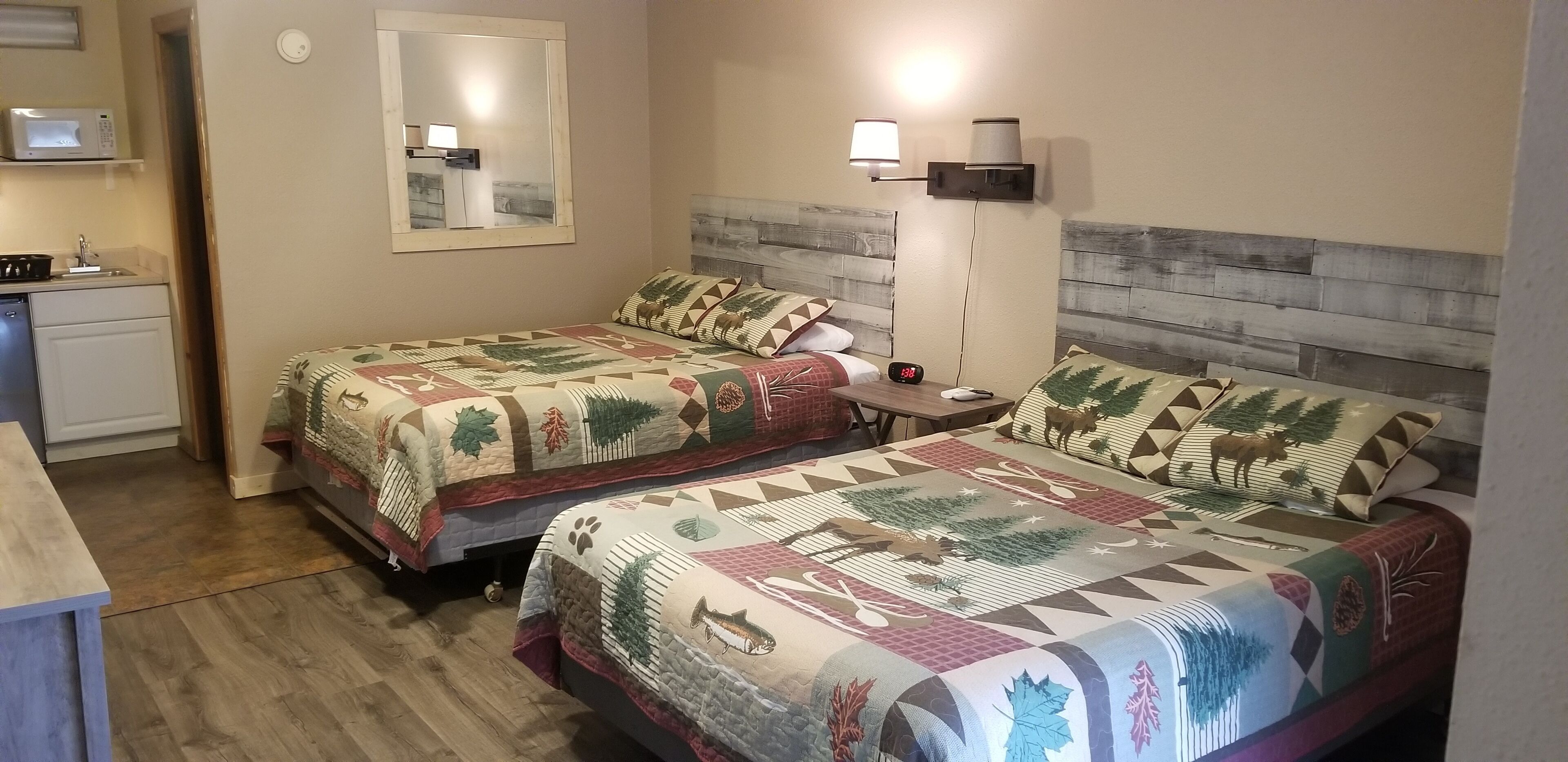 A room with two beds, a small kitchenette, a mirror, and a wall-mounted lamp.
