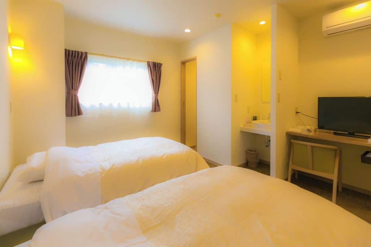 Quarto de hotel com duas camas, uma escrivaninha com televisão, pia e ar condicionado.