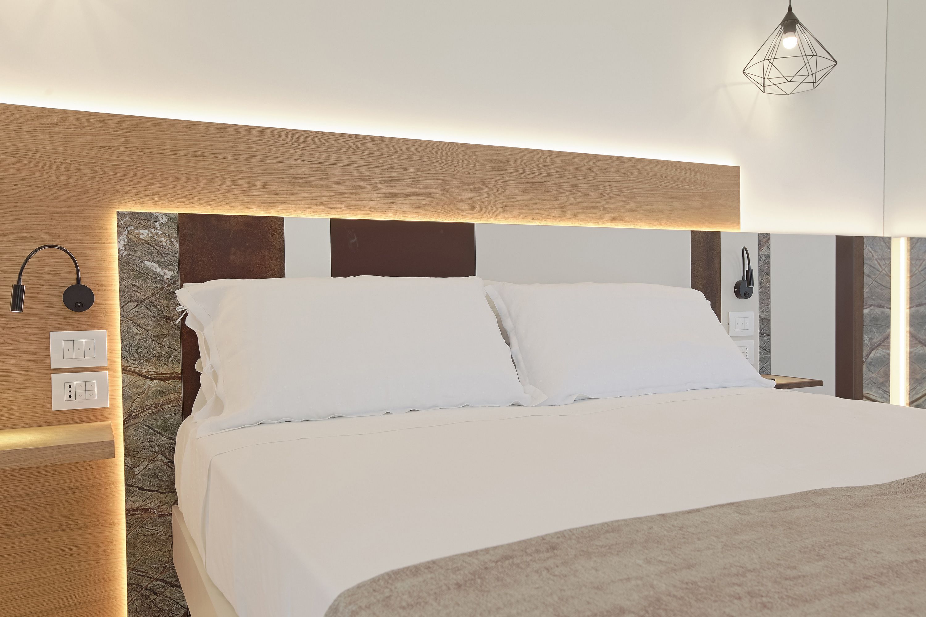 A modern hotel room with a wooden headboard, white bedding, and a stone accent wall.