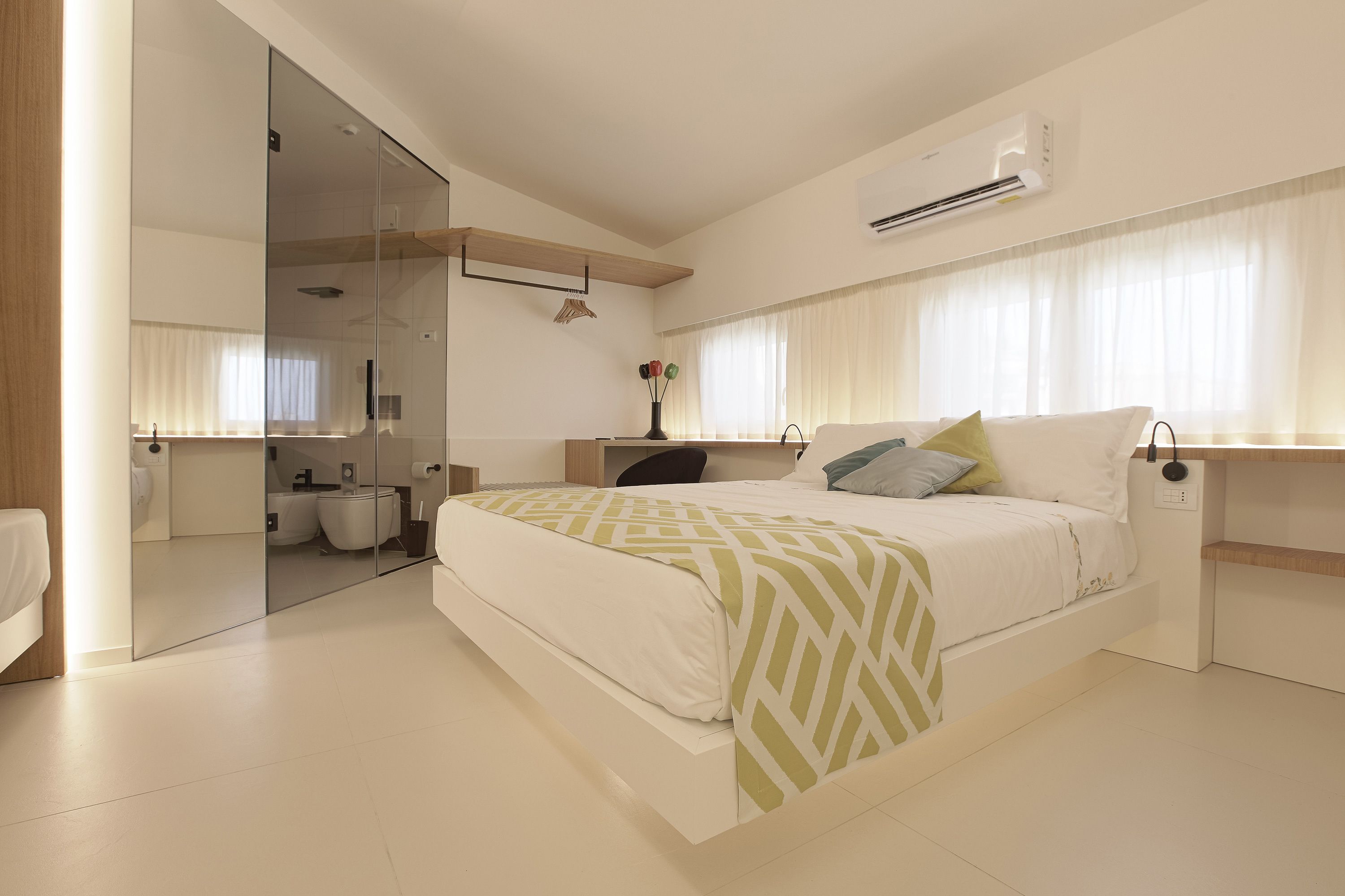 A modern bedroom with a large bed, a bedside table, a chair, a mirror, and a bathroom visible through a glass door.