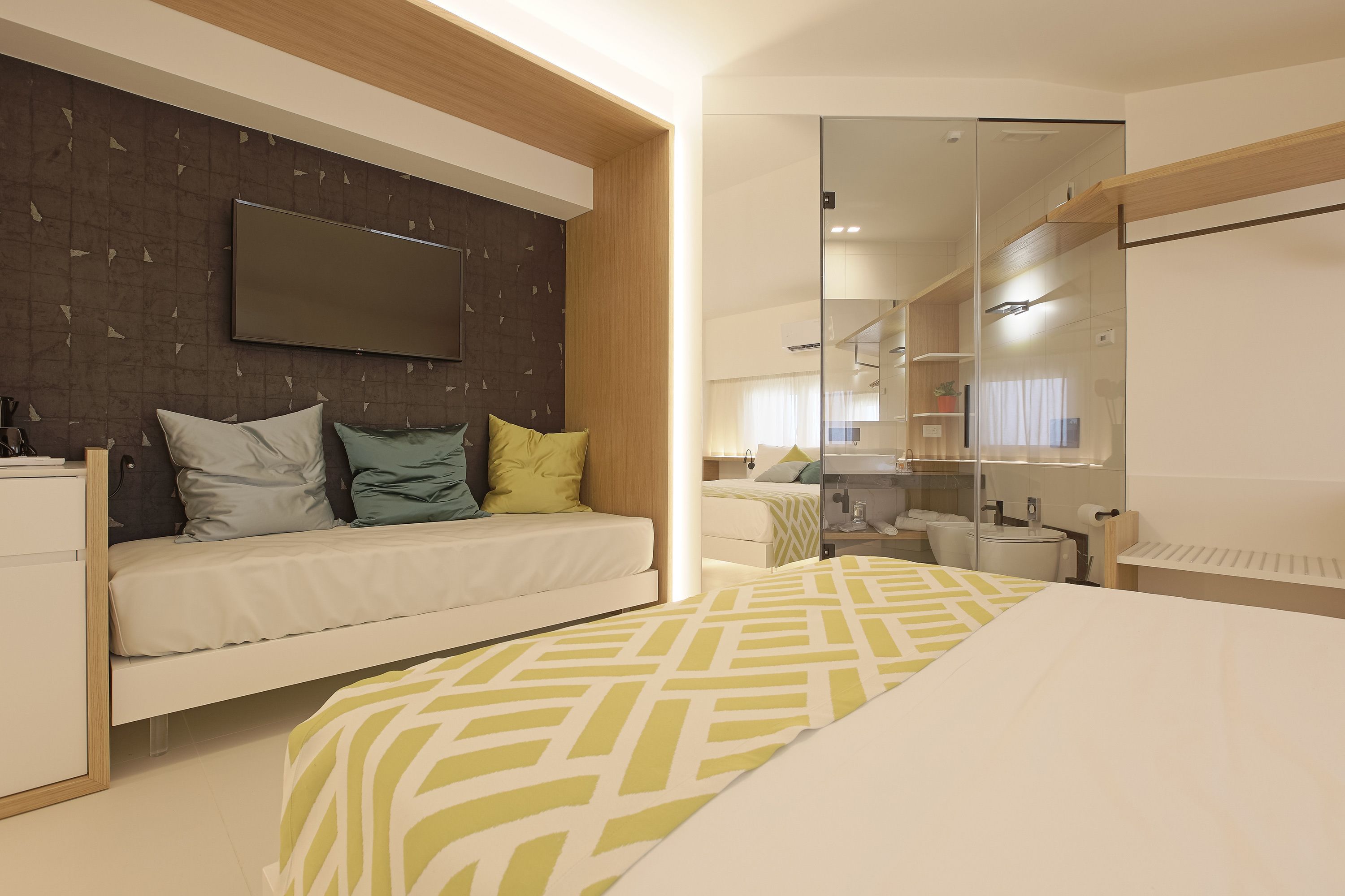 Habitación de hotel moderna con cama, sofá, televisor y baño visible a través de una puerta de vidrio.