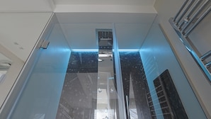 Een moderne lift met glazen deuren en een metalen leuning, die leidt naar een ruimte met hoge plafonds, een ventilatieopening en een lamp.