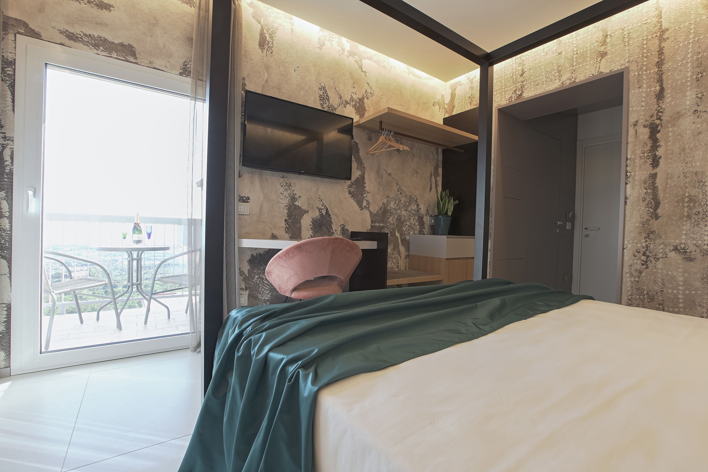 A modern hotel room with a bed, a small desk, a chair, a TV, and a balcony with a table and chairs.