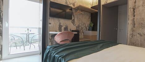 A modern hotel room with a bed, a small desk, a chair, a TV, and a balcony with a table and chairs.