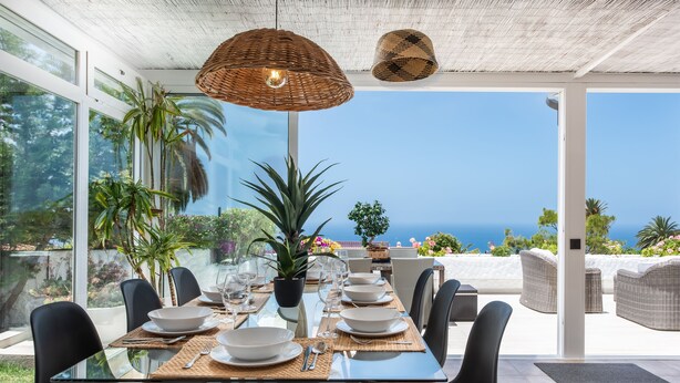 Un comedor con mesa de vidrio, sillas negras y vista al mar.