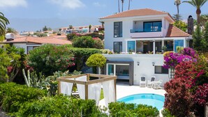 Una casa moderna de dos pisos con piscina, rodeada de exuberante vegetación y palmeras.