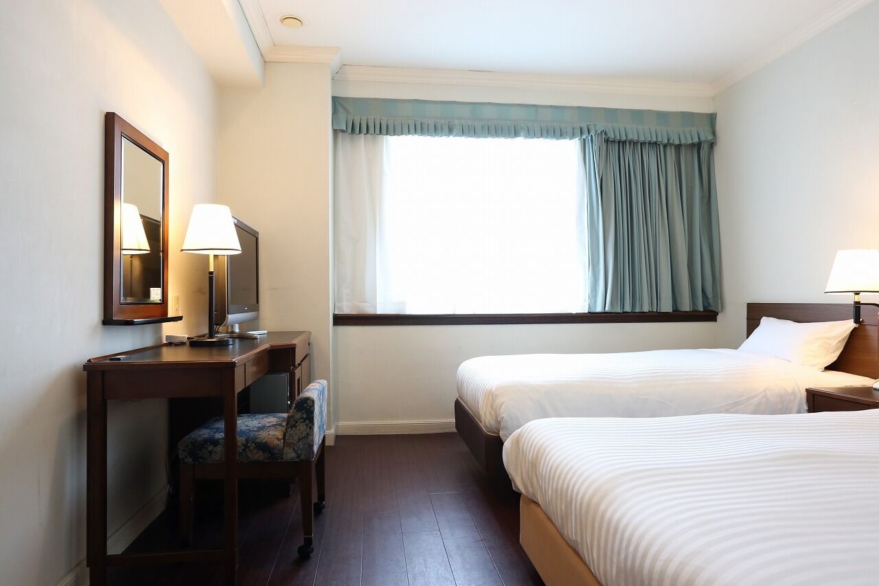 A hotel room with two single beds, a desk with a chair, a mirror, and a window with curtains.