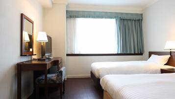 A hotel room with two single beds, a desk with a chair, a mirror, and a window with curtains.