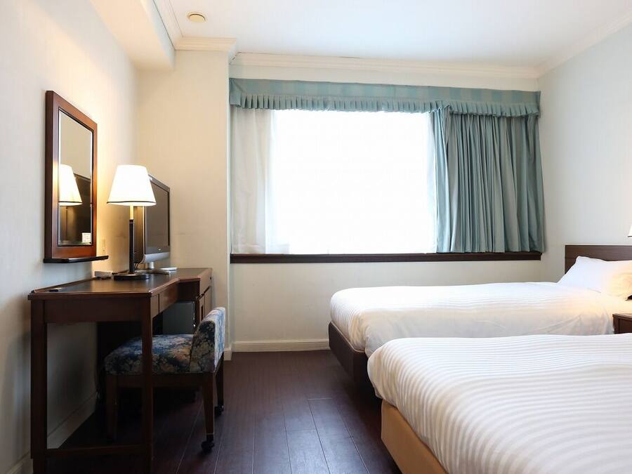 Ein Hotelzimmer mit zwei Einzelbetten, einem Schreibtisch mit Stuhl, einem Spiegel und einem Fenster mit Vorhängen.