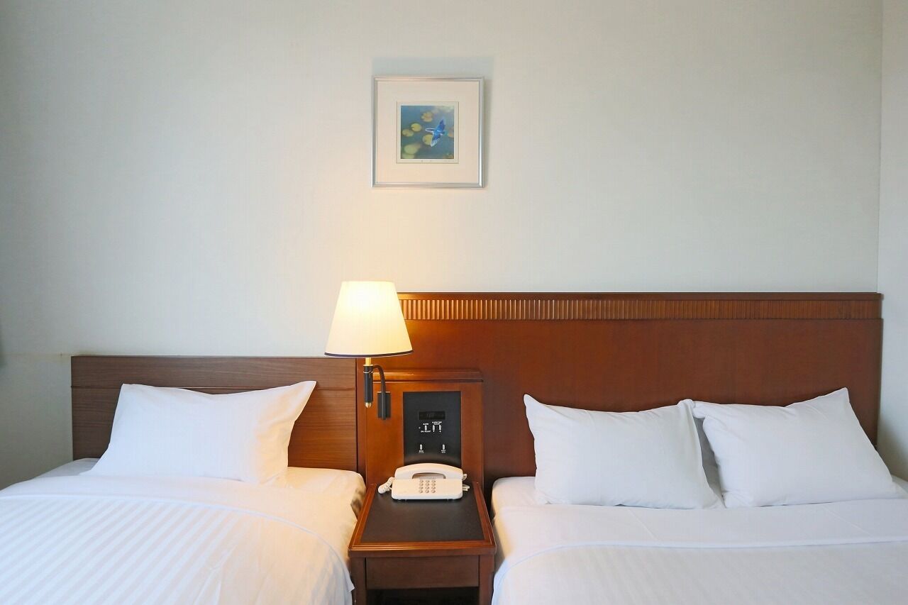 A hotel room with two beds, a bedside table with a lamp, a phone, and a framed picture on the wall.