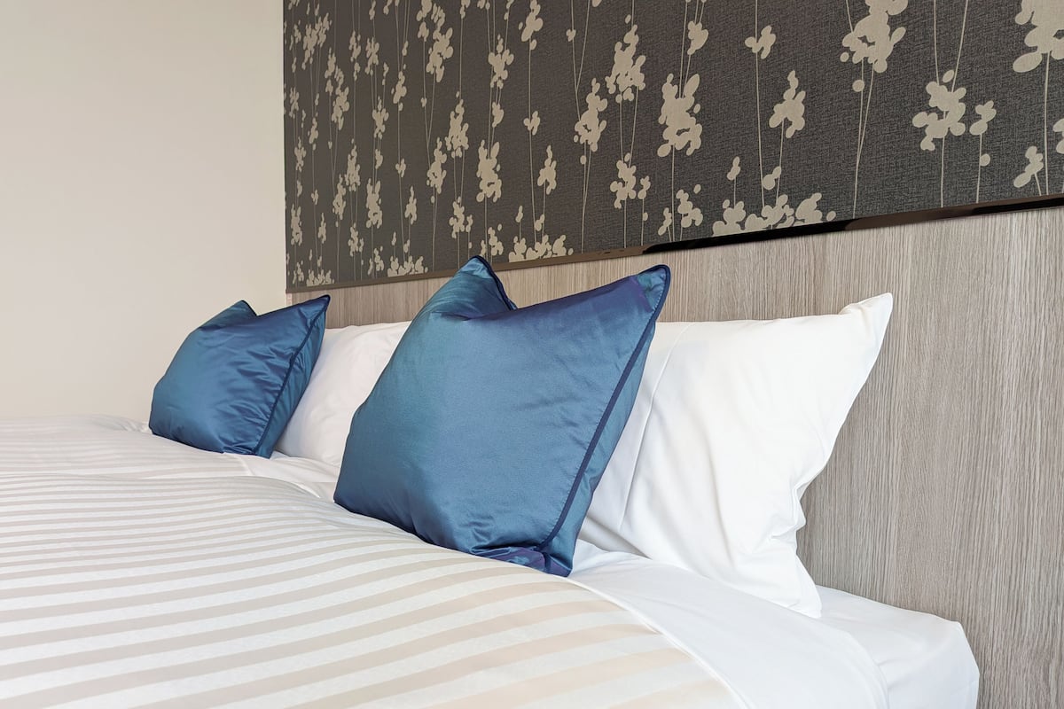 Uma cama bem arrumada com roupa de cama listrada em branco e cinza claro, duas almofadas azuis e uma cabeceira de madeira com padrão floral.