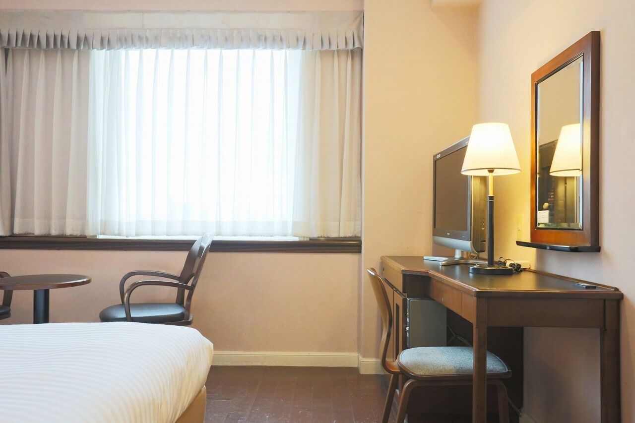 A hotel room with a bed, a desk with a computer and lamp, a chair, and a window with curtains.
