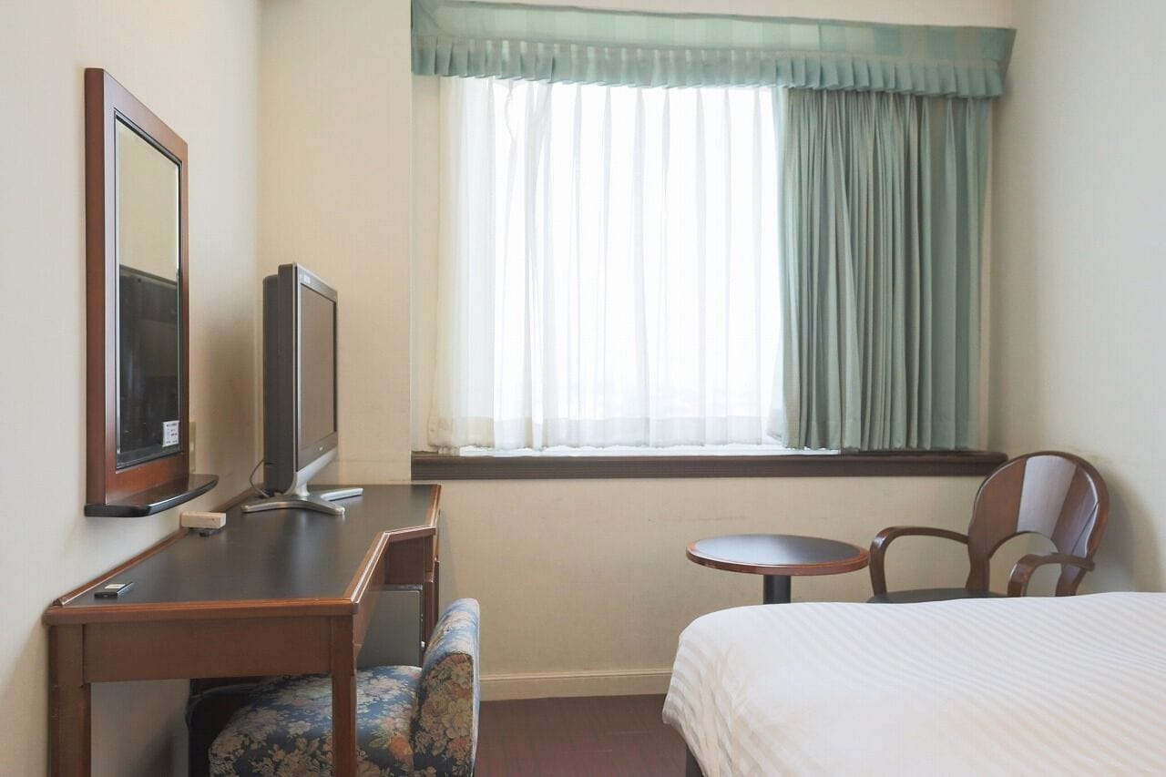 A hotel room with a bed, a desk with a computer, a chair, a small table, and a window with curtains.