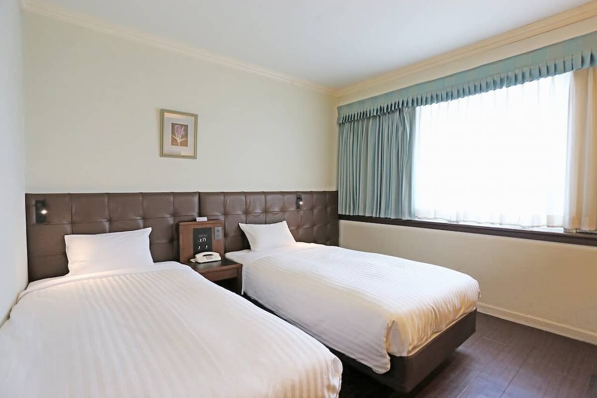 Quarto de hotel com duas camas, cabeceira, mesa de cabeceira com telefone e janela com cortinas.