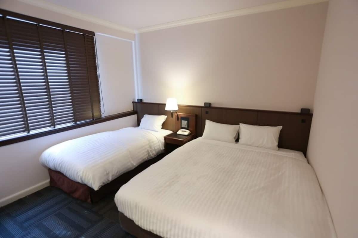 Quarto de hotel com duas camas de solteiro, uma mesa de cabeceira com abajur e uma janela com persianas.