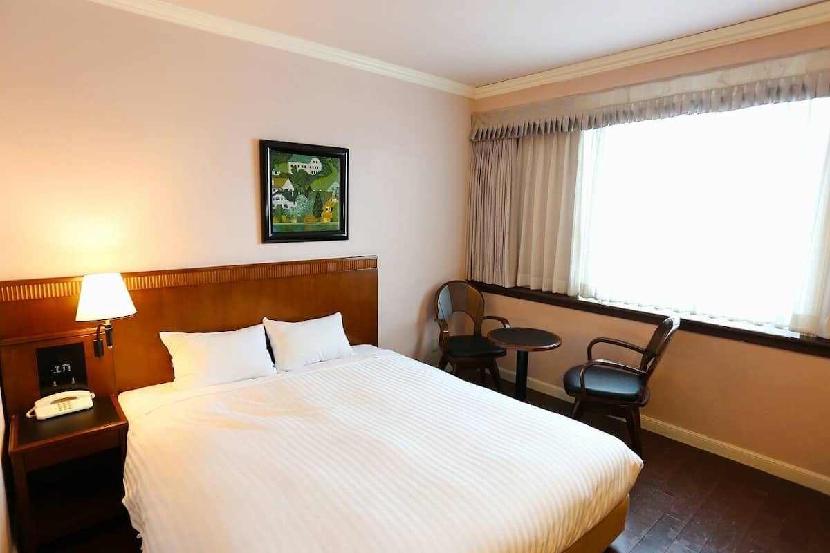 Quarto de hotel com uma cama grande, mesa de cabeceira com abajur, telefone, cadeira, mesinha e janela com cortinas.