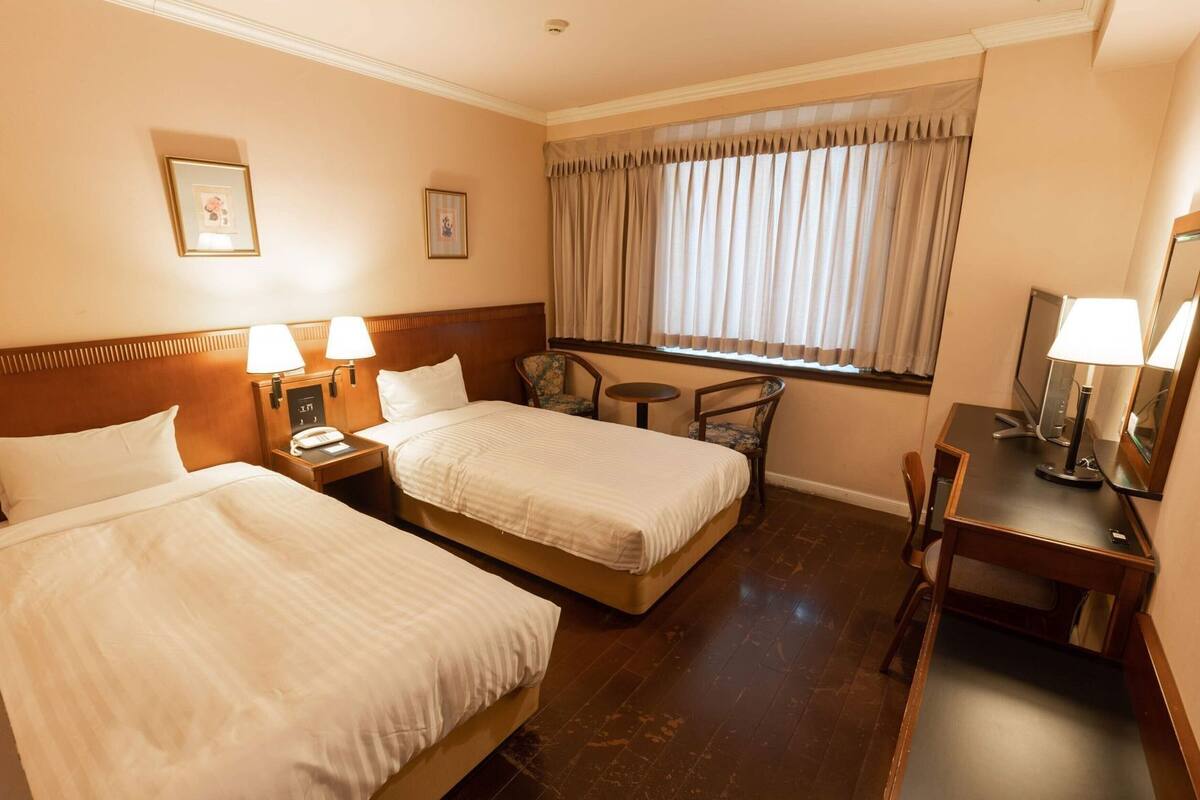 A hotel room with two beds, a desk, a chair, a small table, and a window with curtains.