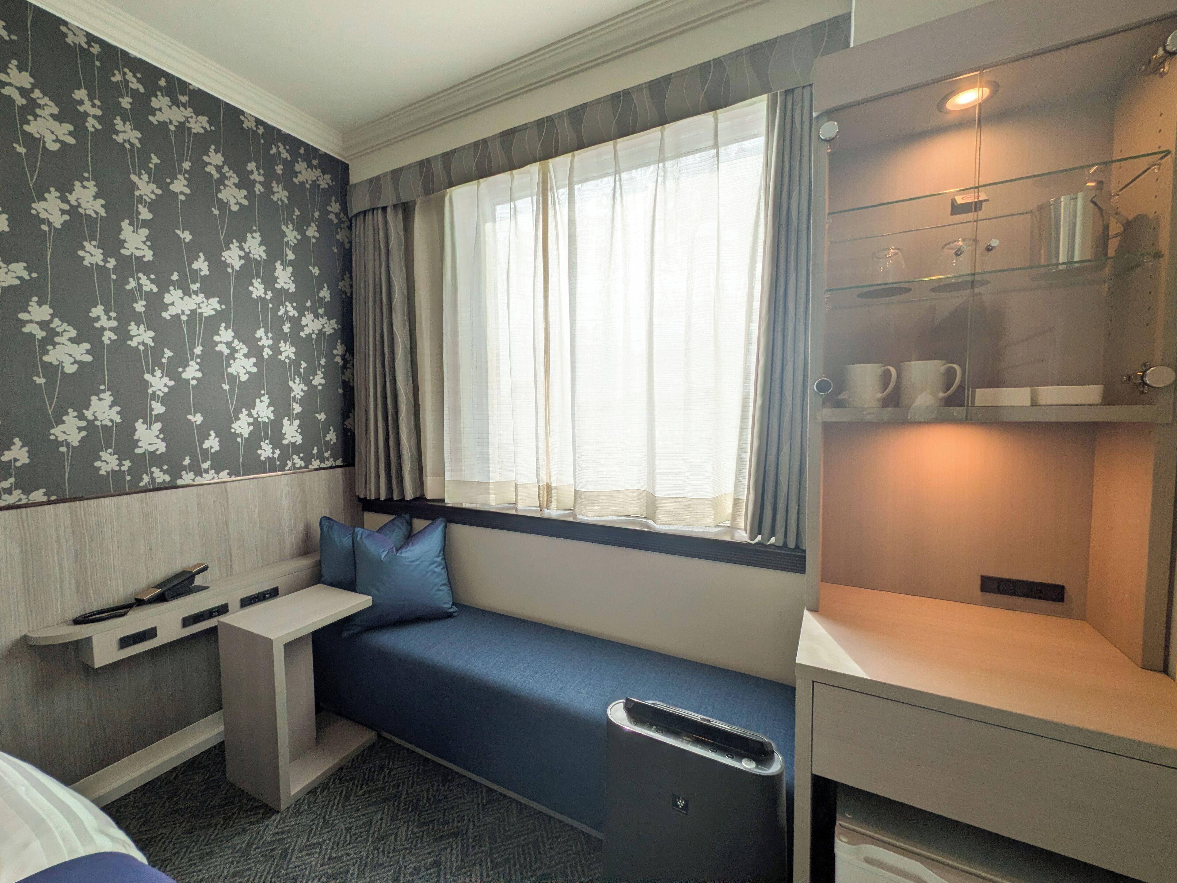 A compact hotel room with a built-in bench along the window, a small desk, and a shelf with cups and a kettle.