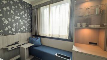 A compact hotel room with a built-in bench along the window, a small desk, and a shelf with cups and a kettle.