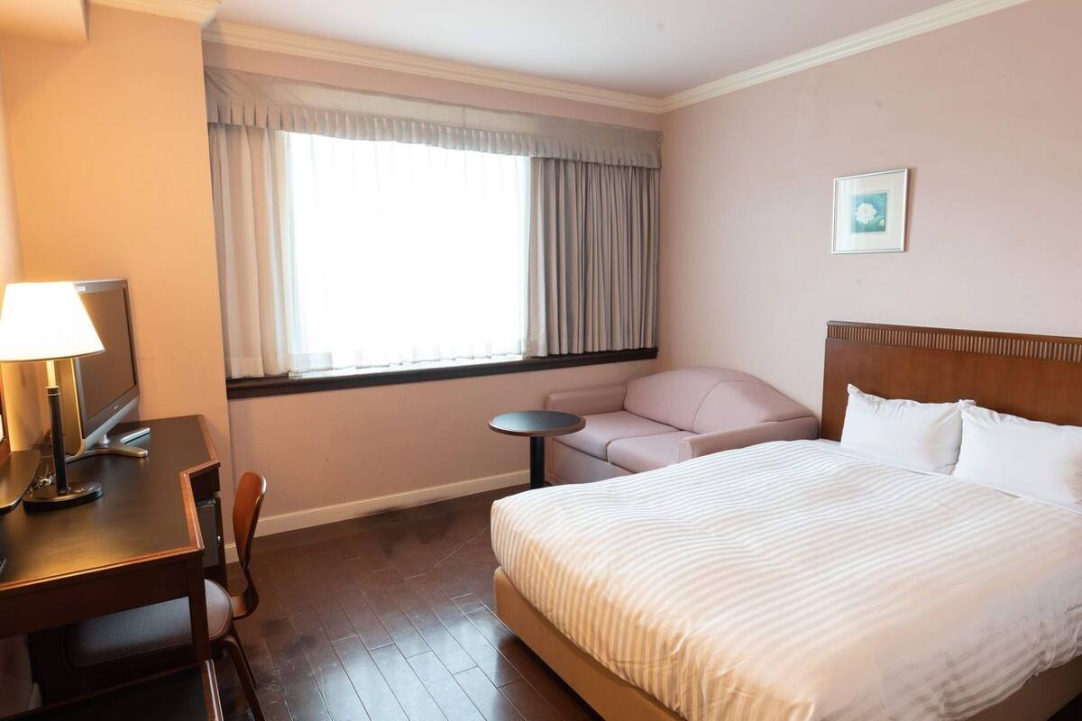 A hotel room with a bed, a desk with a lamp, a chair, a small table, and a window with curtains.