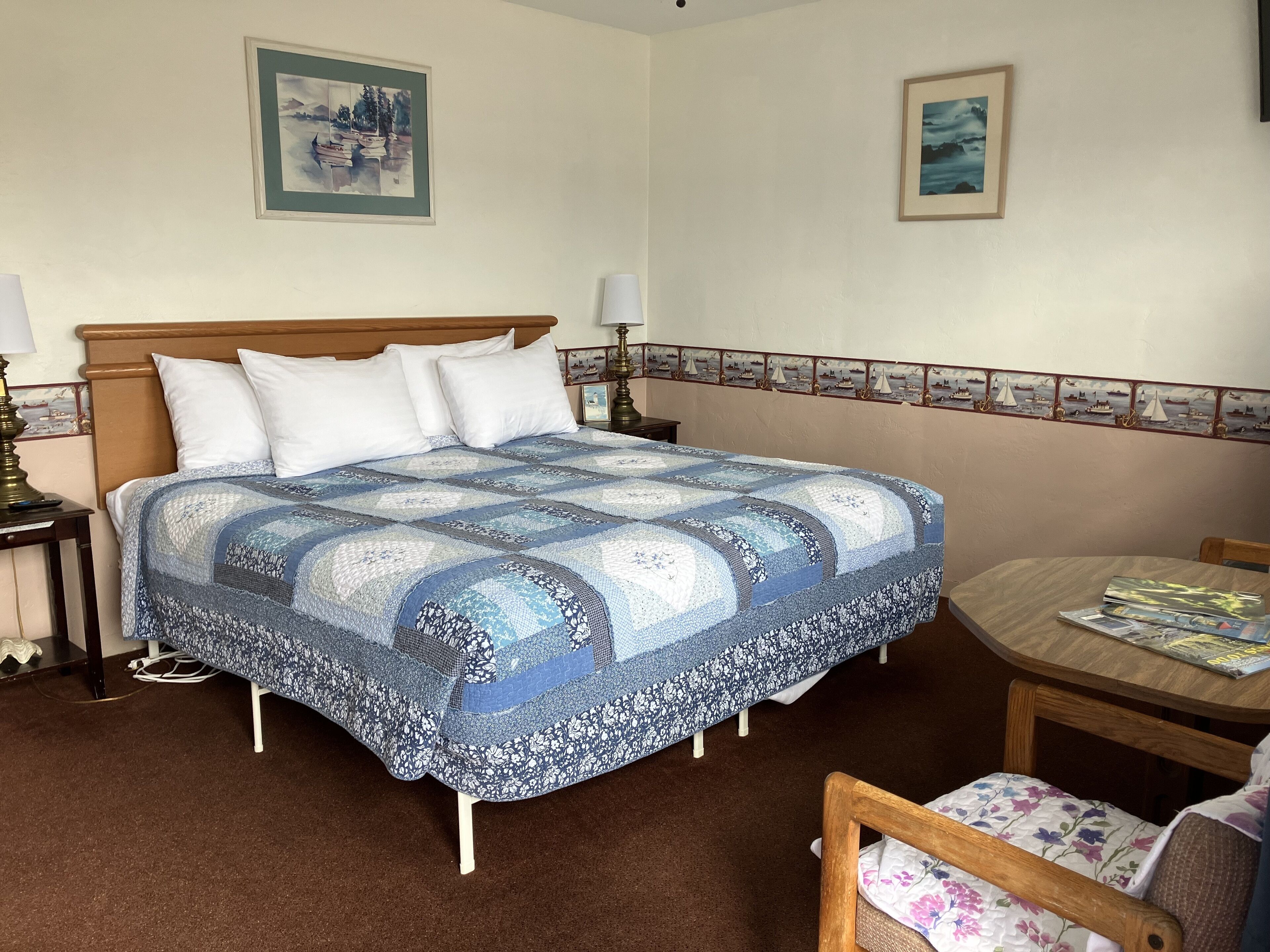 A neatly made bed with a blue and white quilt, a wooden headboard, and two bedside lamps. There is a small table with a chair beside the bed, and a framed picture on the wall above the bed.
