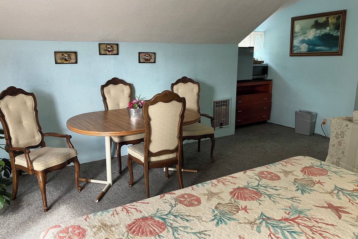A room with a dining table, four chairs, a bed with a floral bedspread, a dresser, a radiator, and a painting on the wall.
