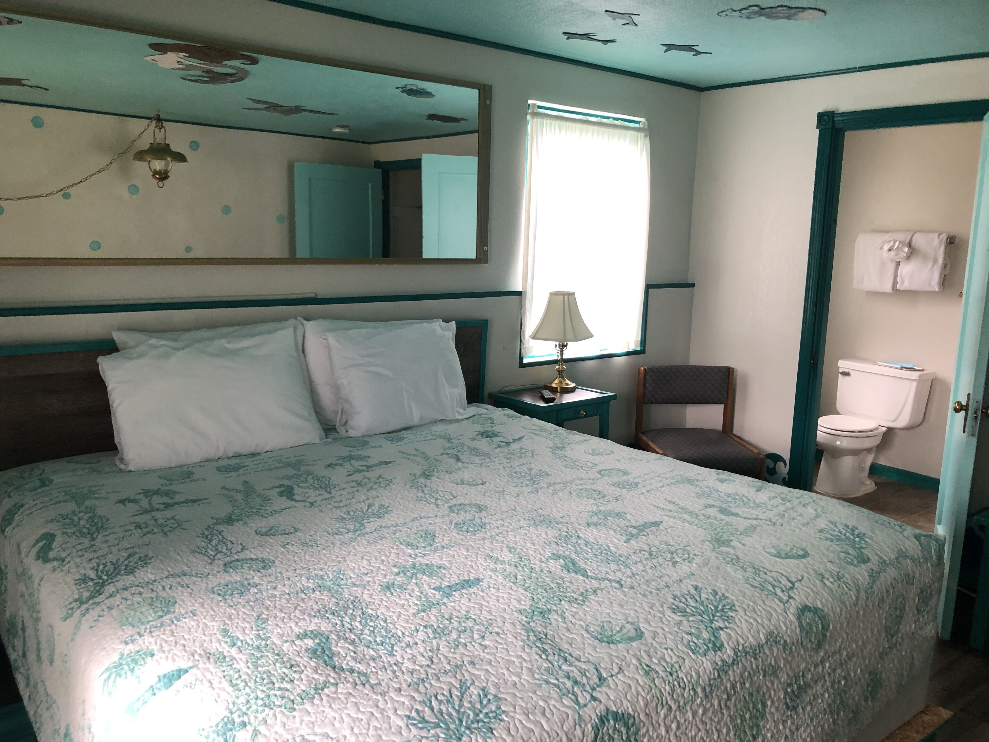 A hotel room with a bed, a chair, a nightstand with a lamp, a window with curtains, a toilet, and a mirror on the wall.