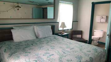 A hotel room with a bed, a chair, a nightstand with a lamp, a window with curtains, a toilet, and a mirror on the wall.