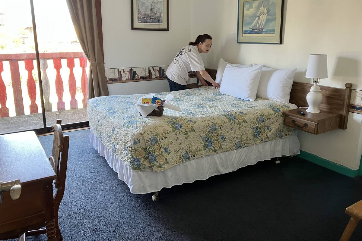 A hotel room with a bed, a nightstand, a lamp, a chair, a table, and a window with curtains.