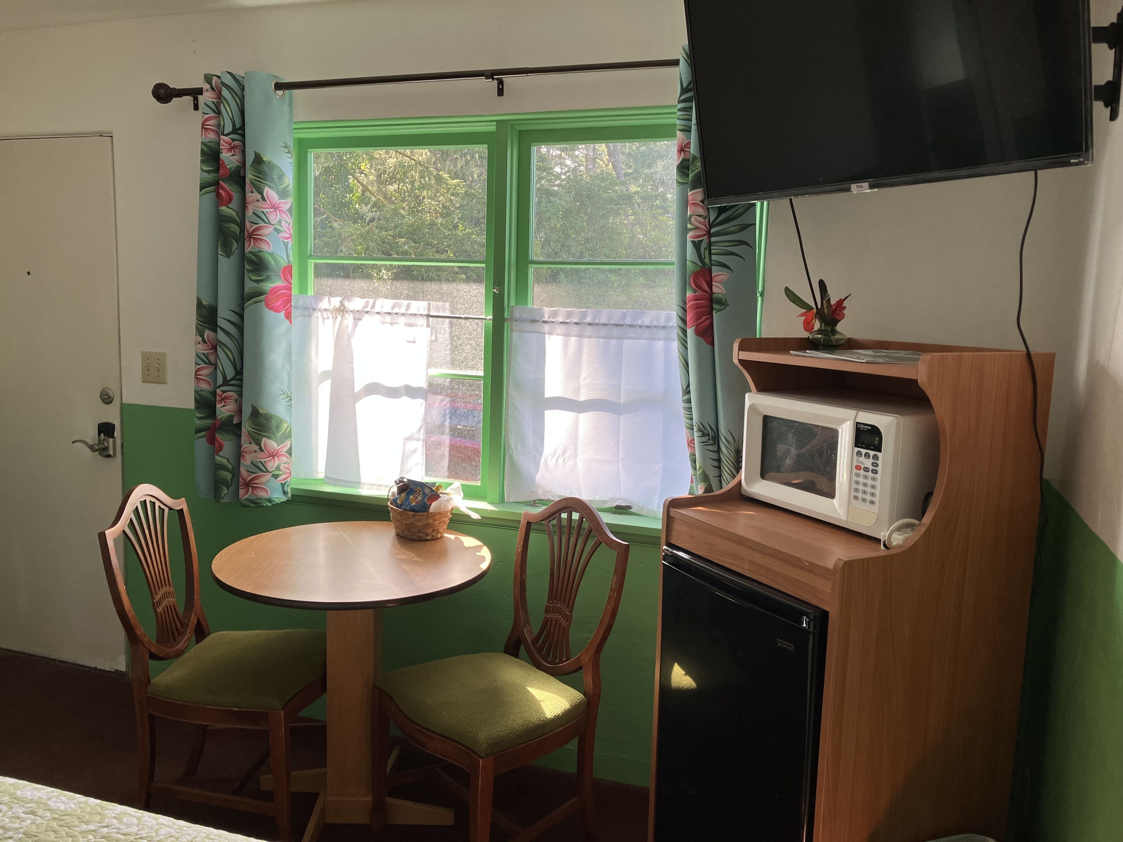 A small room with a green window, a wooden table with two chairs, a microwave, and a television mounted on the wall.