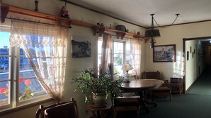 A dining area with a round table and chairs, a potted plant, and a view of the street through a window with lace curtains.