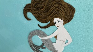 A mermaid cutout with brown hair and a grey tail against a teal background.