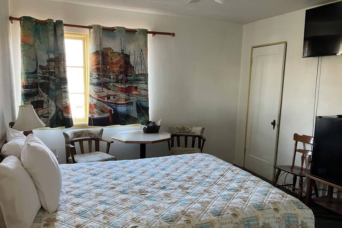 A hotel room with a bed, a small table, a chair, a TV, and a window with curtains.