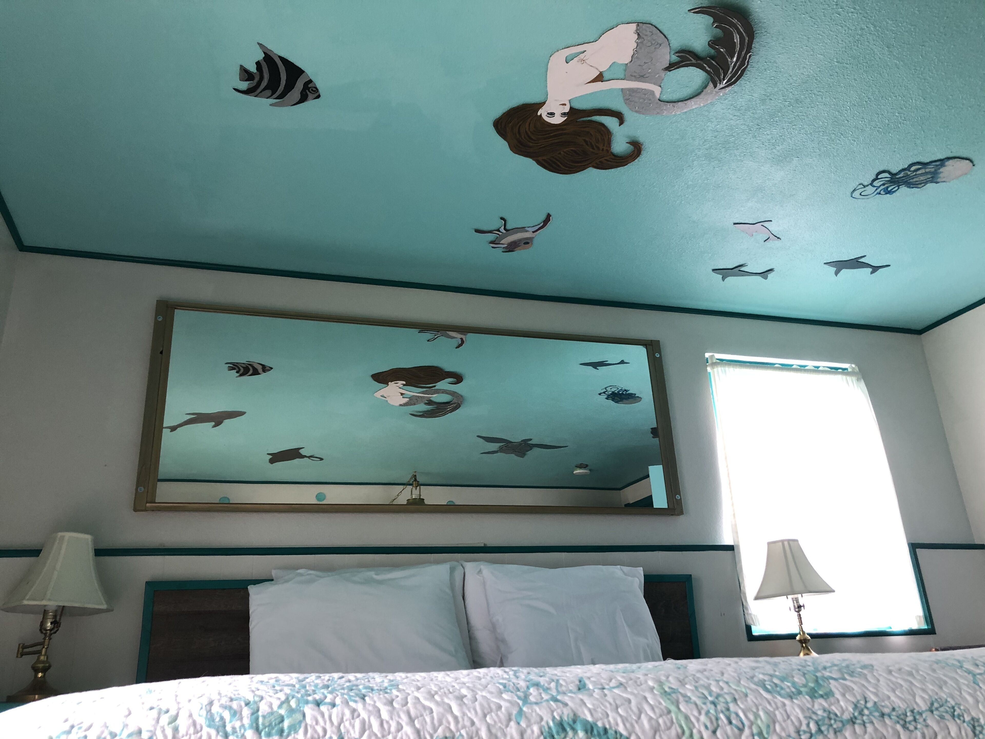 A hotel room with a teal ceiling decorated with mermaid and fish decals, a large mirror, a bed with white bedding, and a window with a view.