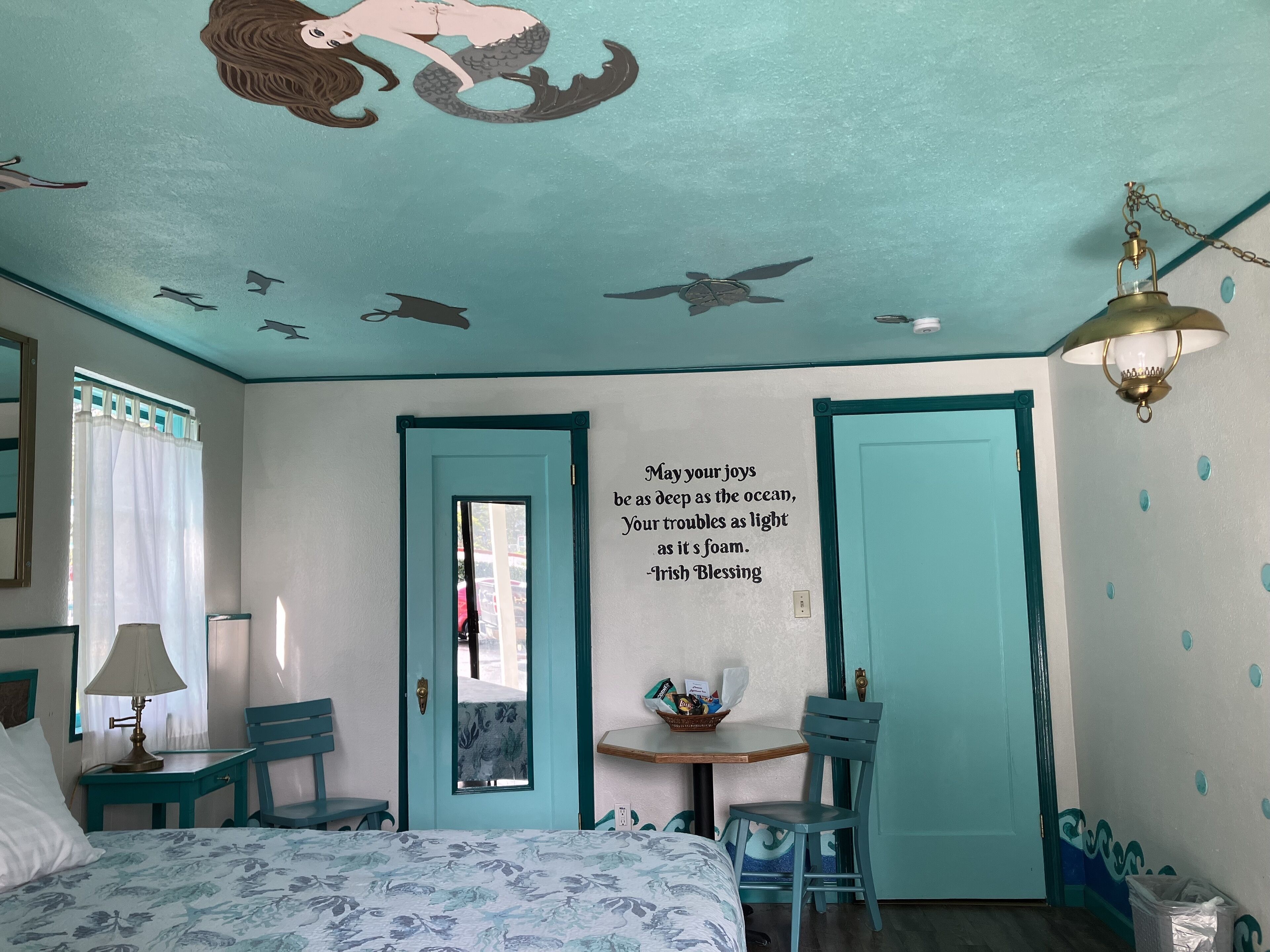 Una habitación con cama, escritorio, silla, puerta y pared con un mural de sirena y una cita.