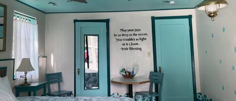 Una habitación con cama, escritorio, silla, puerta y pared con un mural de sirena y una cita.