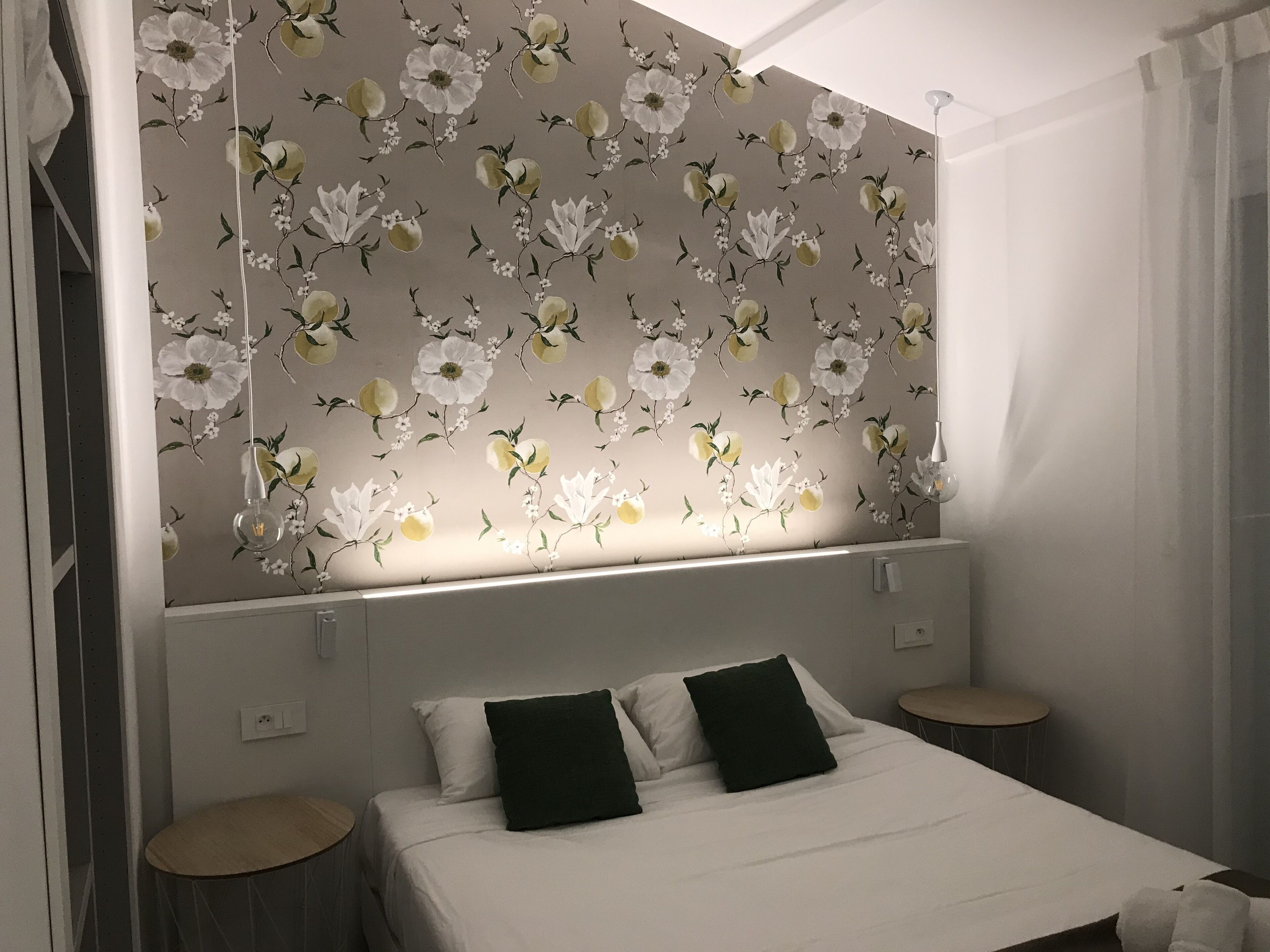 Une chambre à coucher avec un papier peint à motifs floraux, un lit avec des oreillers blancs et vert foncé, et deux tables de chevet.