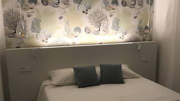A neatly made bed with a white and gray color scheme, a headboard with a nature-themed wallpaper, and a small bedside table with a lamp.