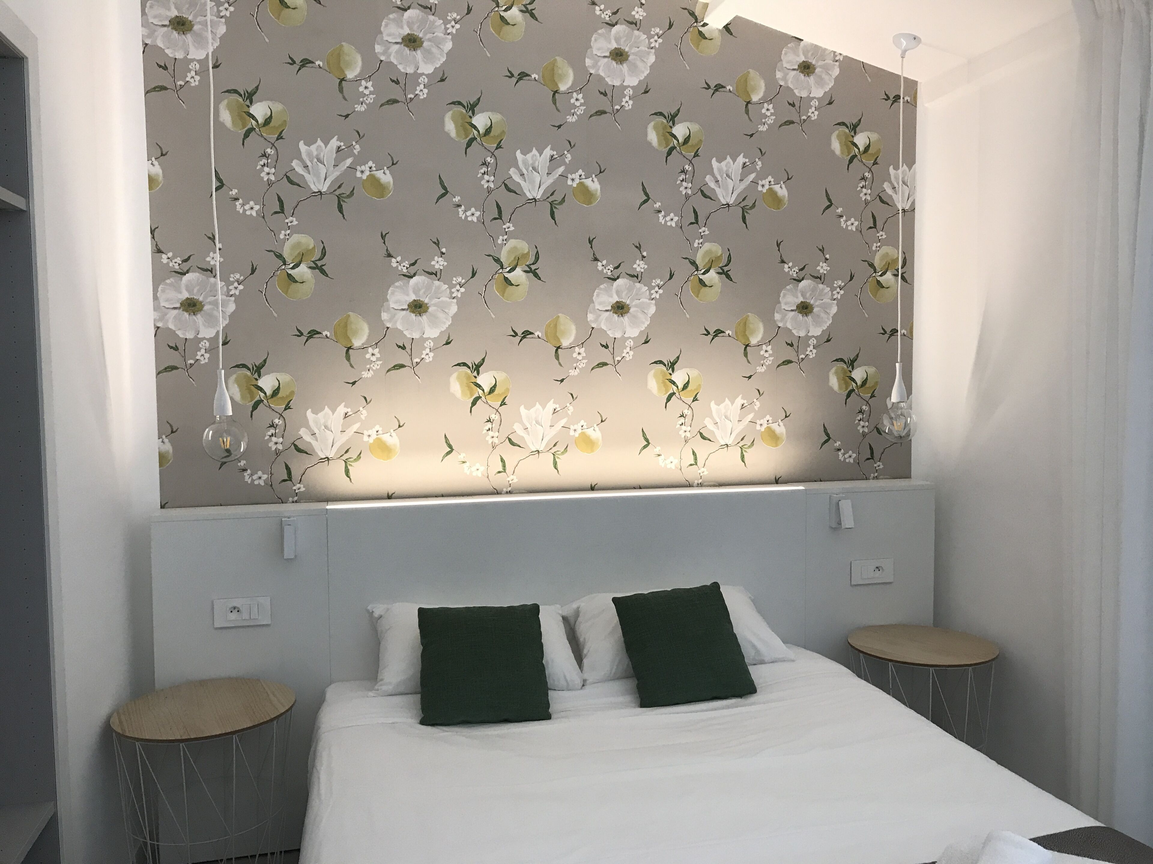 A bedroom with a floral wallpaper, a bed with white and green pillows, and two round side tables.