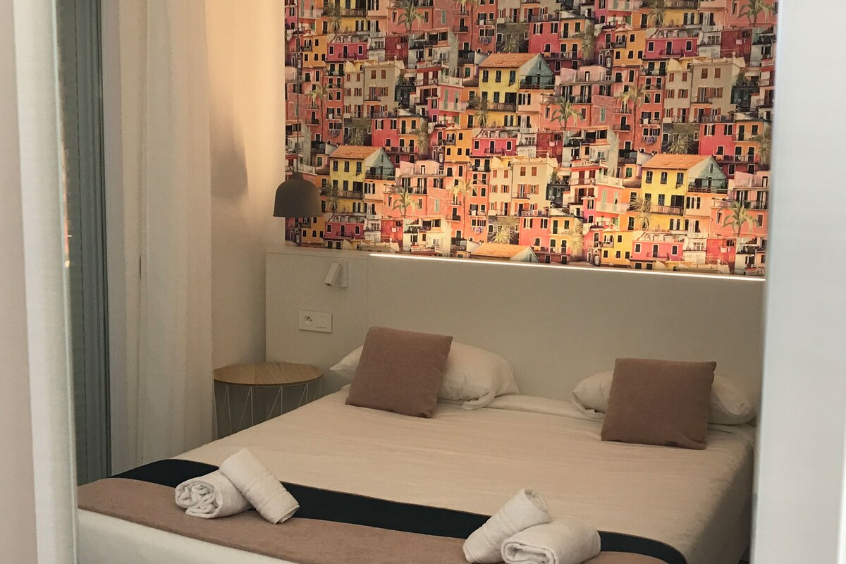 A hotel room with a bed, pillows, a bedside lamp, and a wall with colorful building patterns.