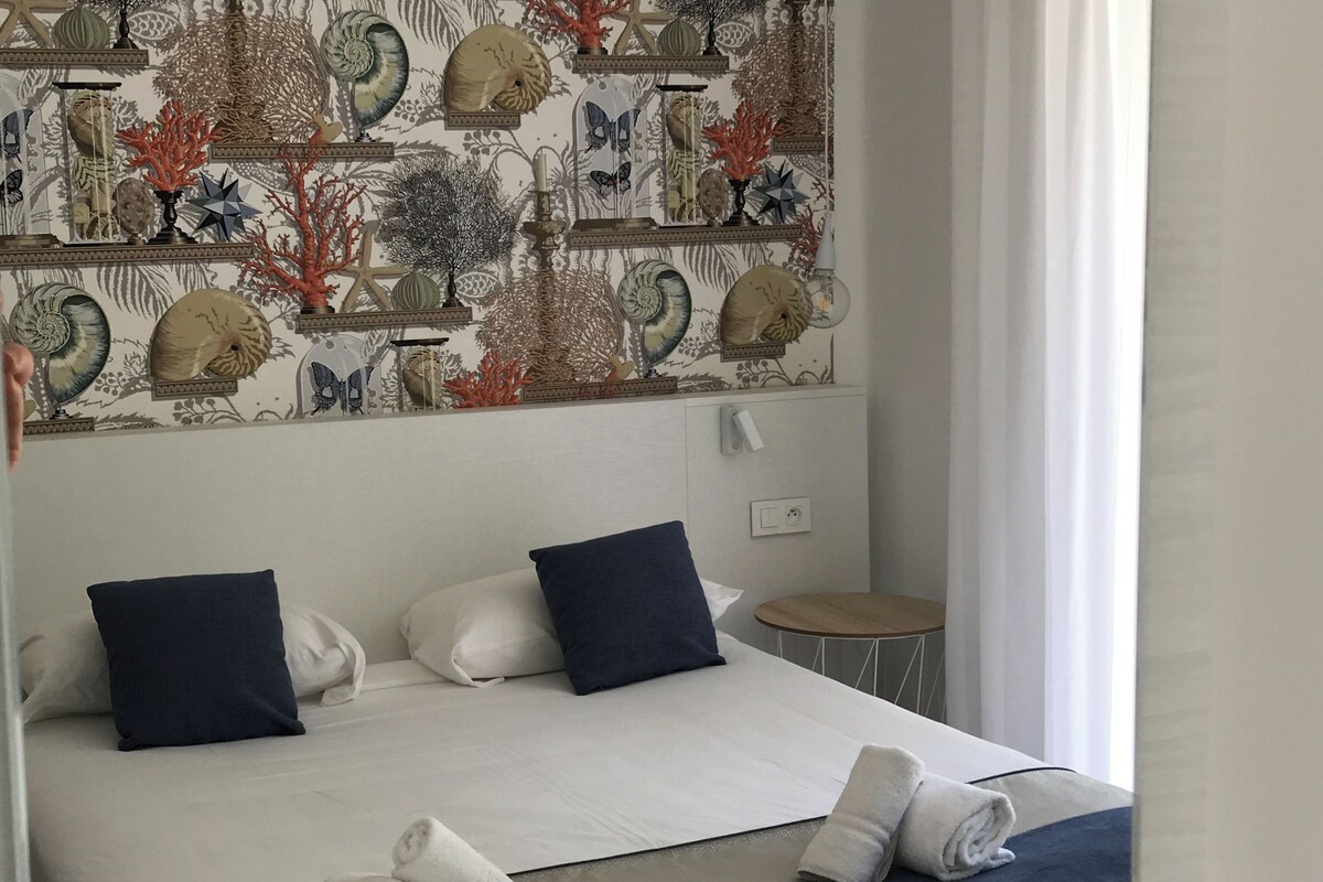 Una cama bien hecha con sábanas blancas y almohadas azul marino, una mesita redonda a un lado y una pared con un diseño decorativo.