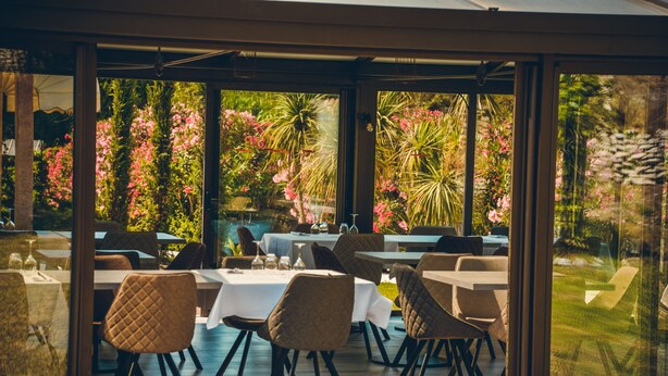 An outdoor dining area with tables and chairs, surrounded by glass walls offering views of a garden with pink flowers and greenery. - Prea Gianca (Bonifacio)