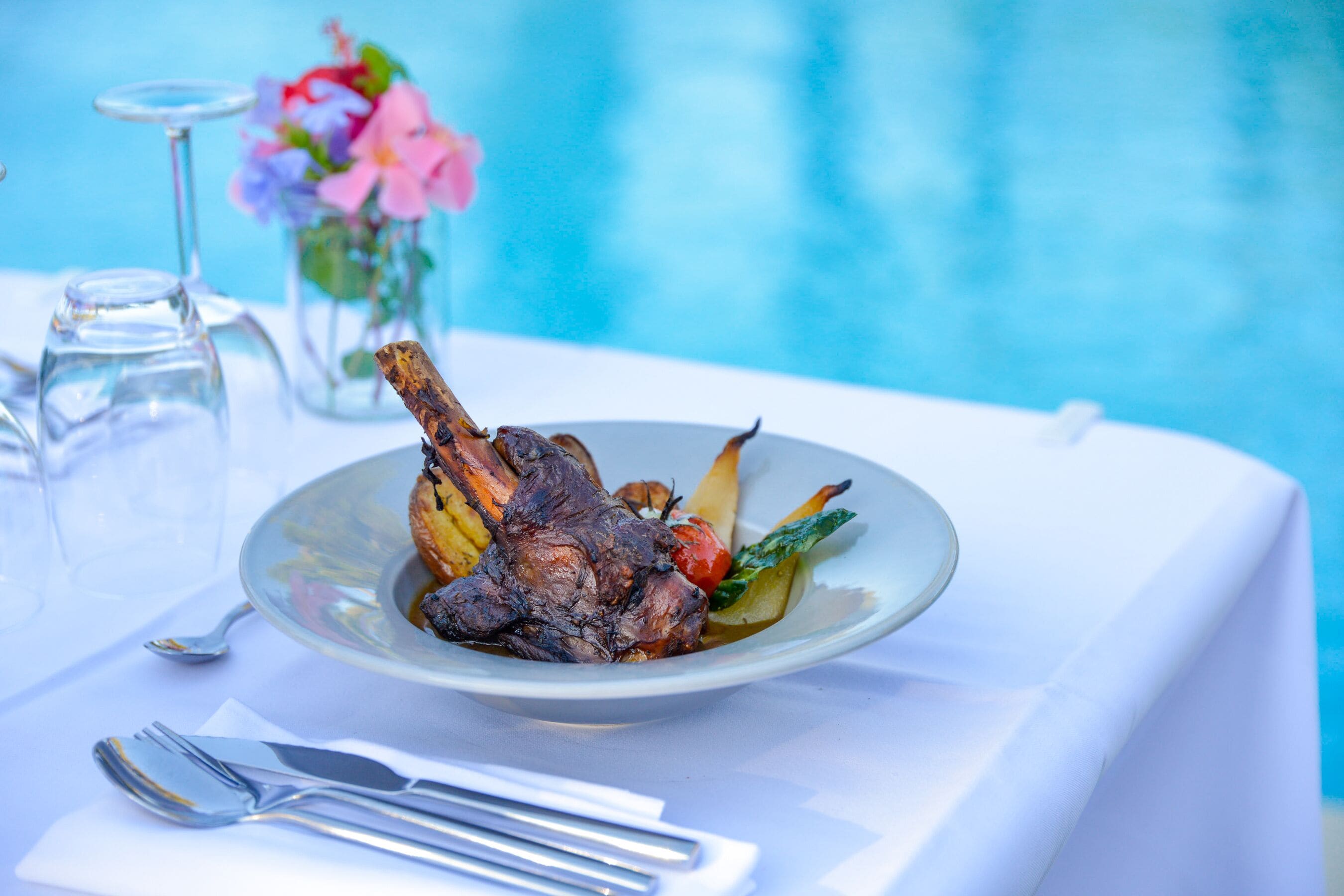 A plate of food with a lamb shank, vegetables, and a glass of water on a table by a pool.