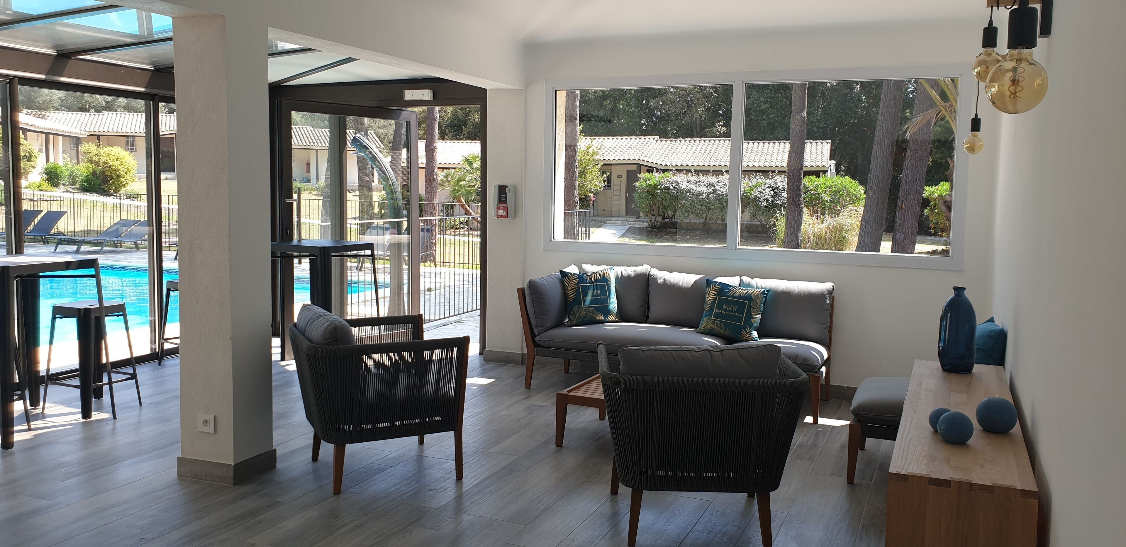 A modern living room with a sofa, chairs, and a wooden table. A large window offers a view of a pool and outdoor area.