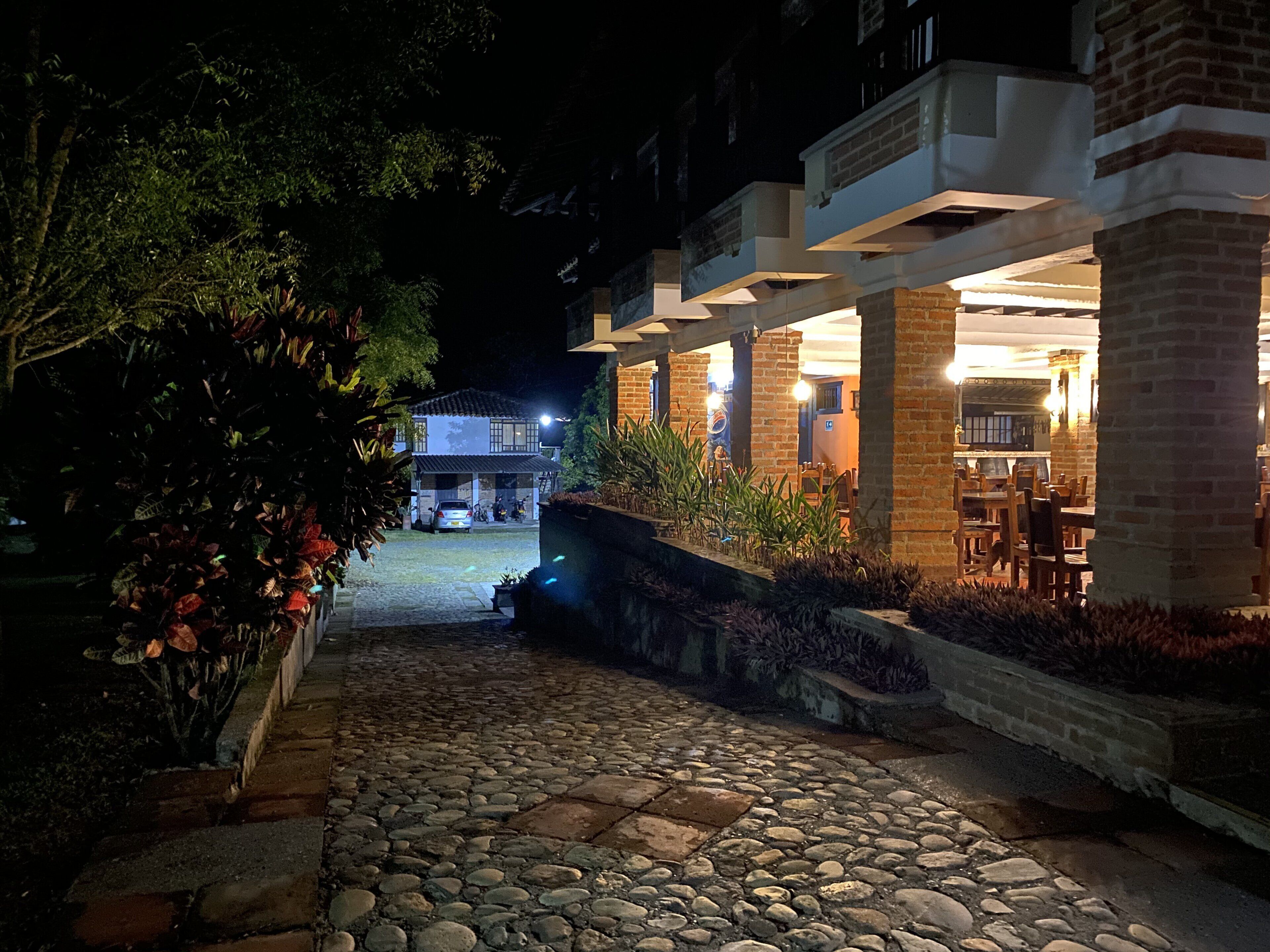 A nighttime view of a courtyard with a stone pathway leading to a building with a brick facade and outdoor seating area.